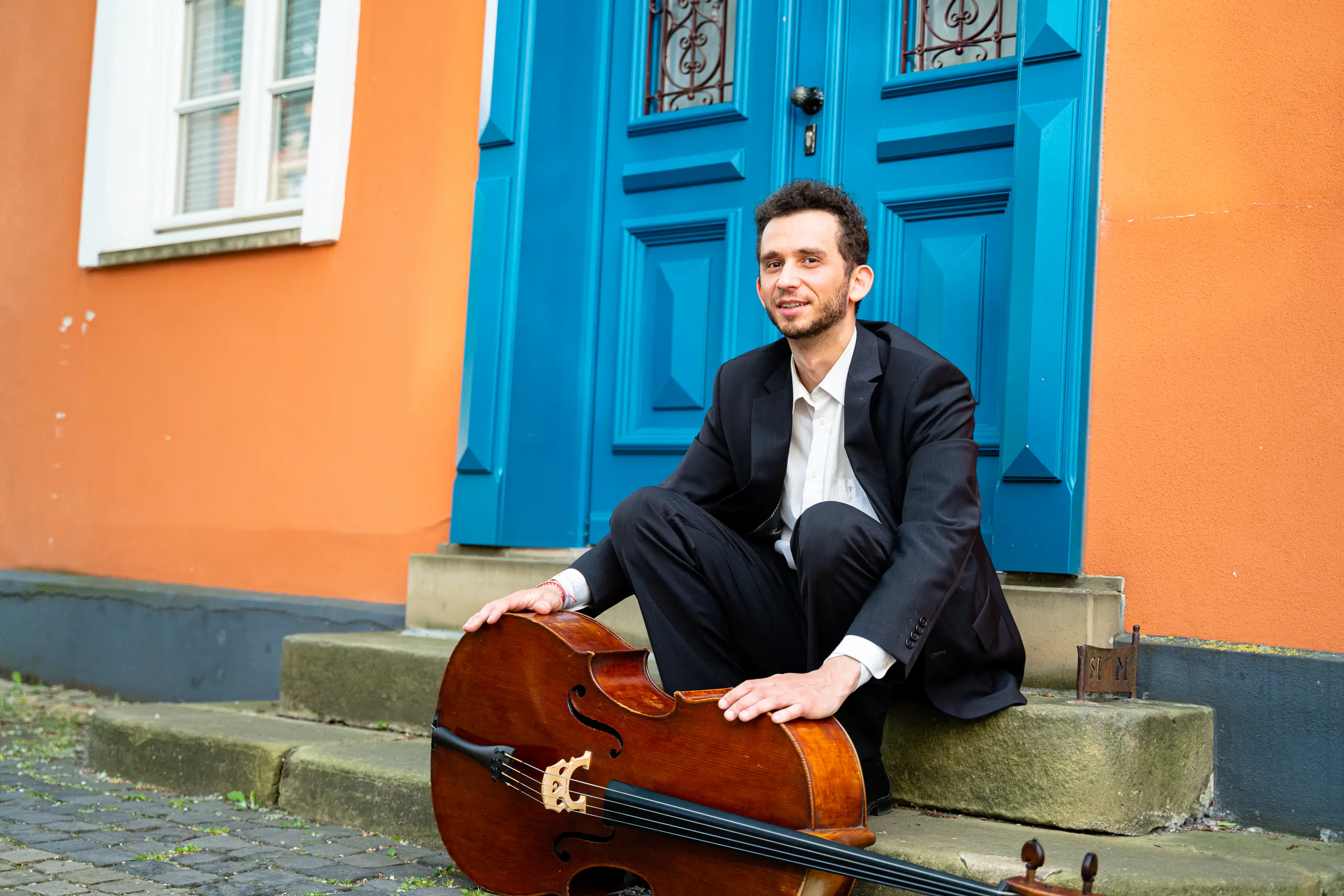 Ventsislav Harkov sitting with his cello on the steps in front of a blue door