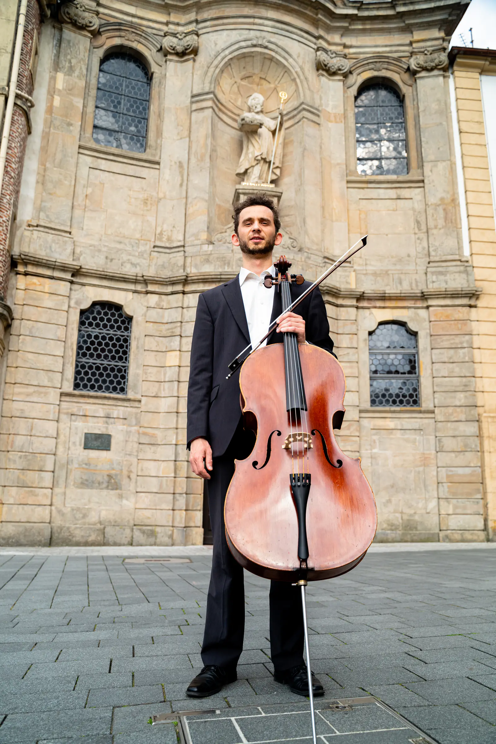 Ventsislav Harkov standing with his cello in front of a Baroque church facade