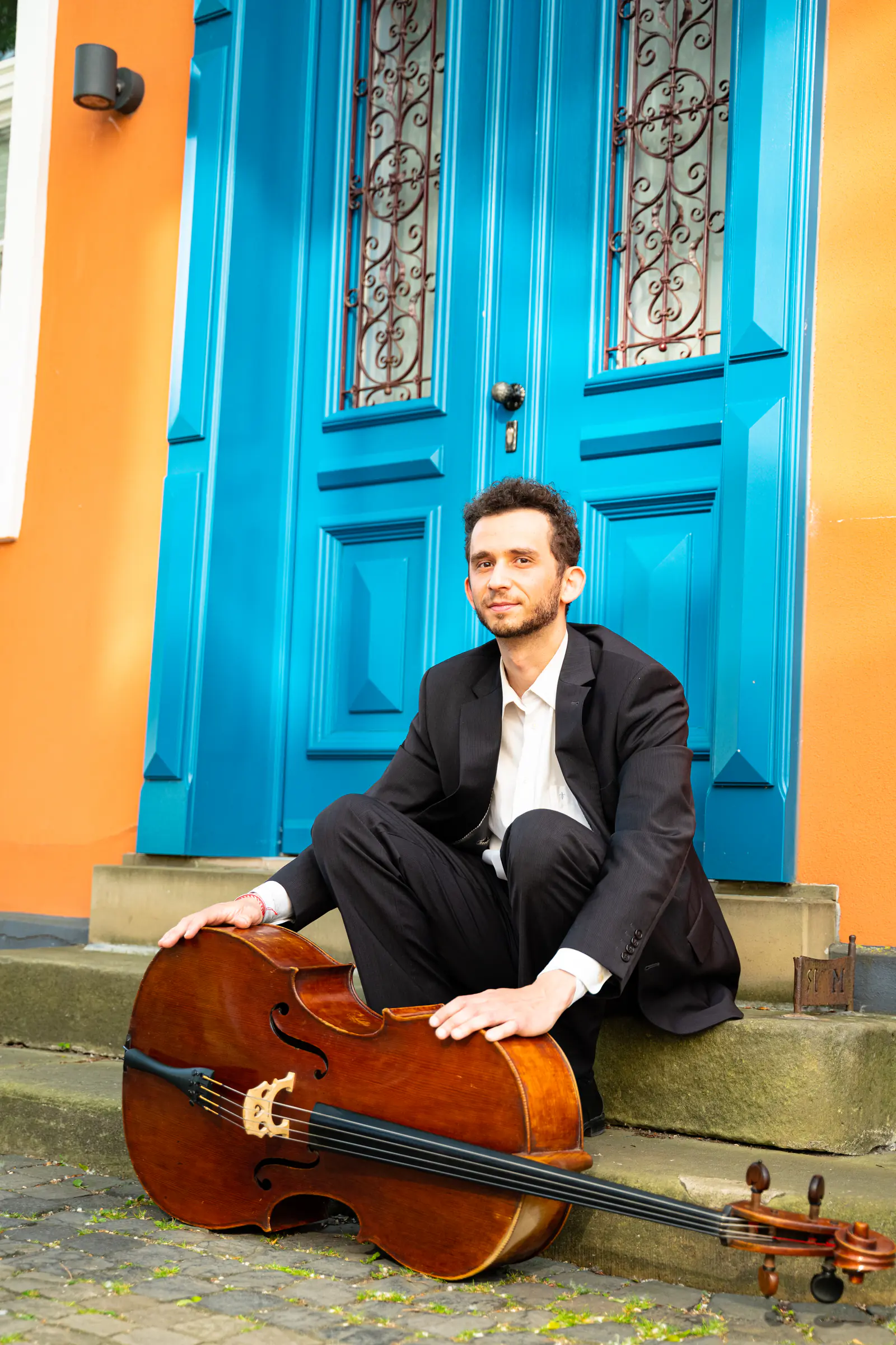 Ventsislav Harkov sitting on the steps in front of a blue door, his hand on his cello