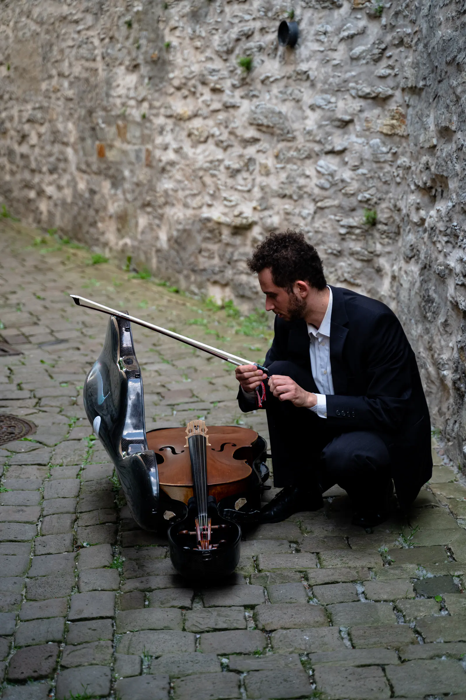 Ventsislav Harkov crouching by his open cello case and preparing his bow beside an old stone wall