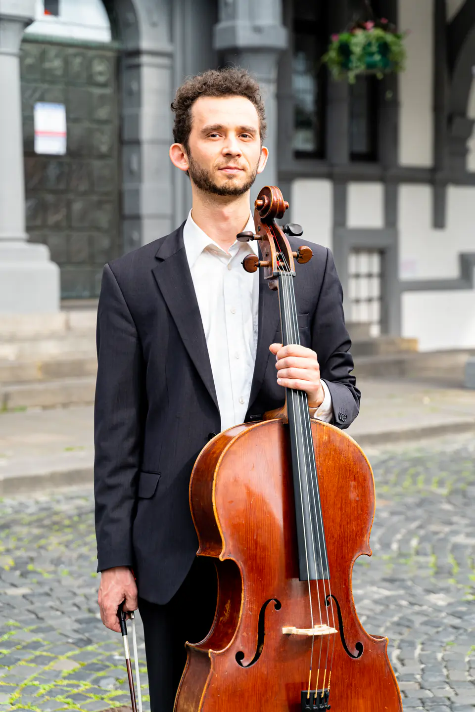 Ventsislav Harkov standing with his cello on a cobbled square in front of a historic building