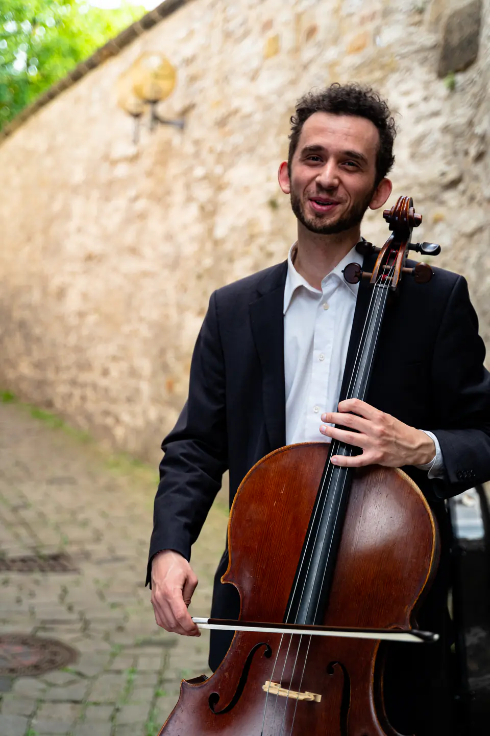 Ventsislav Harkov laughing, with his cello and bow, in a lane along a stone wall