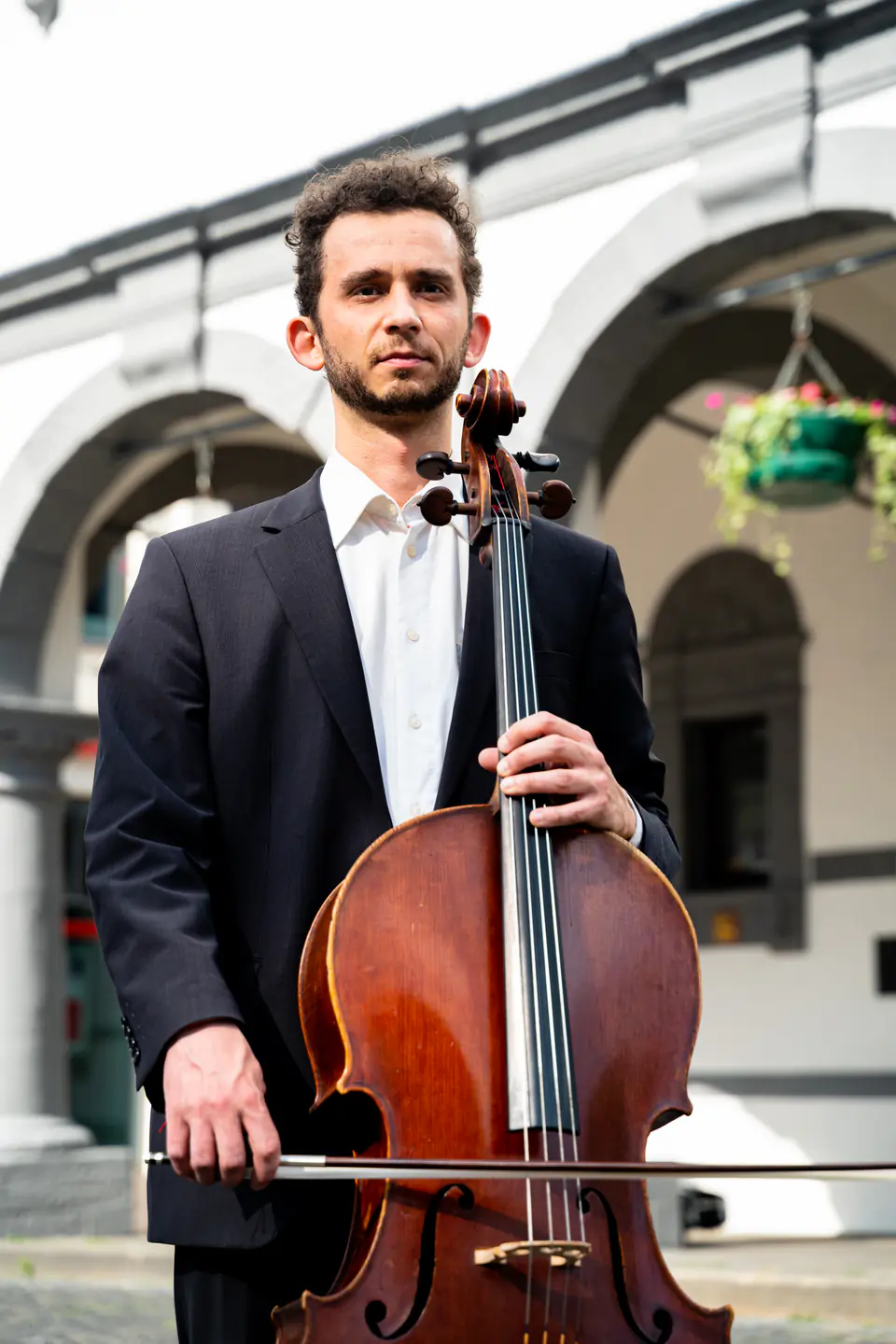 Ventsislav Harkov holding his cello and bow in front of the arches of a white building