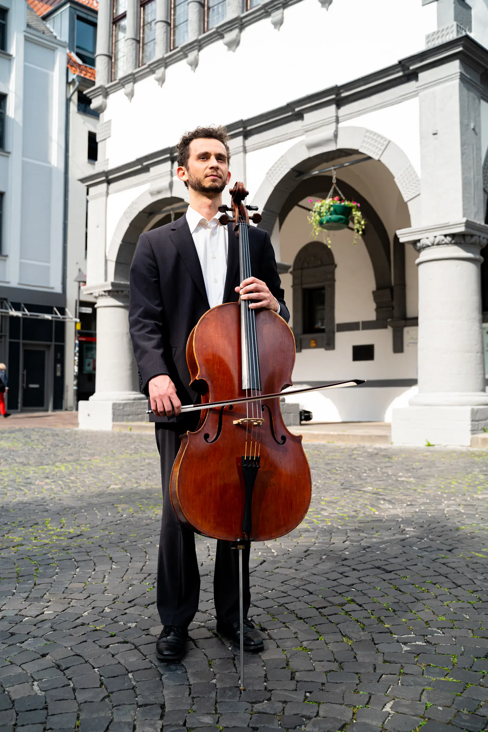 Ventsislav Harkov with his cello and bow in front of a white arcaded building