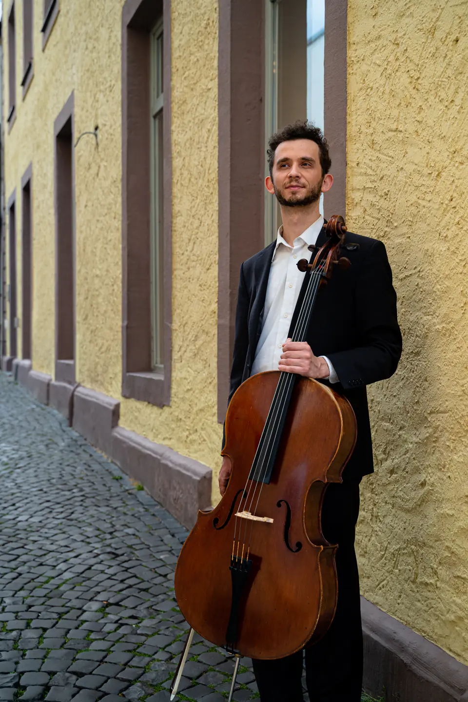Ventsislav Harkov with his cello by a yellow wall, looking into the distance